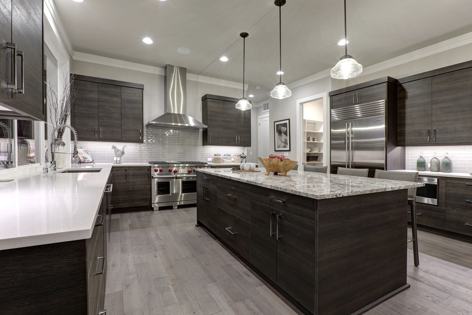 Modern kitchen with dark wood cabinets, stainless steel appliances, a large island with granite countertop, pendant lighting, and light wood flooring.