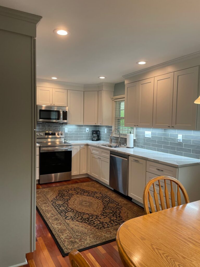Modern kitchen remodeling with white cabinets, stainless steel appliances, gray subway tile backsplash, under-cabinet lighting, and a patterned rug. Wooden dining table and chairs in the foreground.