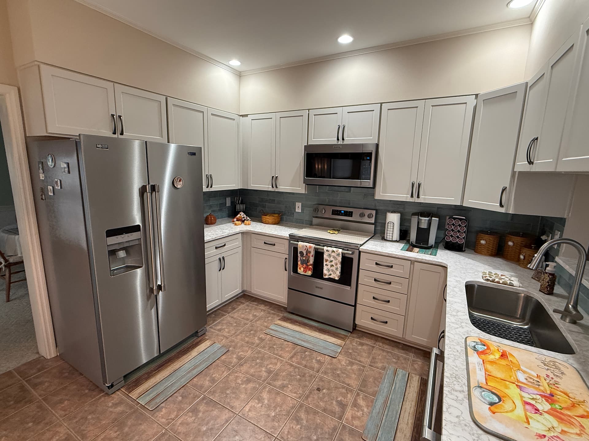 Modern kitchen featuring stainless steel appliances, white cabinets enhanced by kitchen refacing, marble countertops, and a tiled floor. Various kitchen accessories and small appliances line the counters.