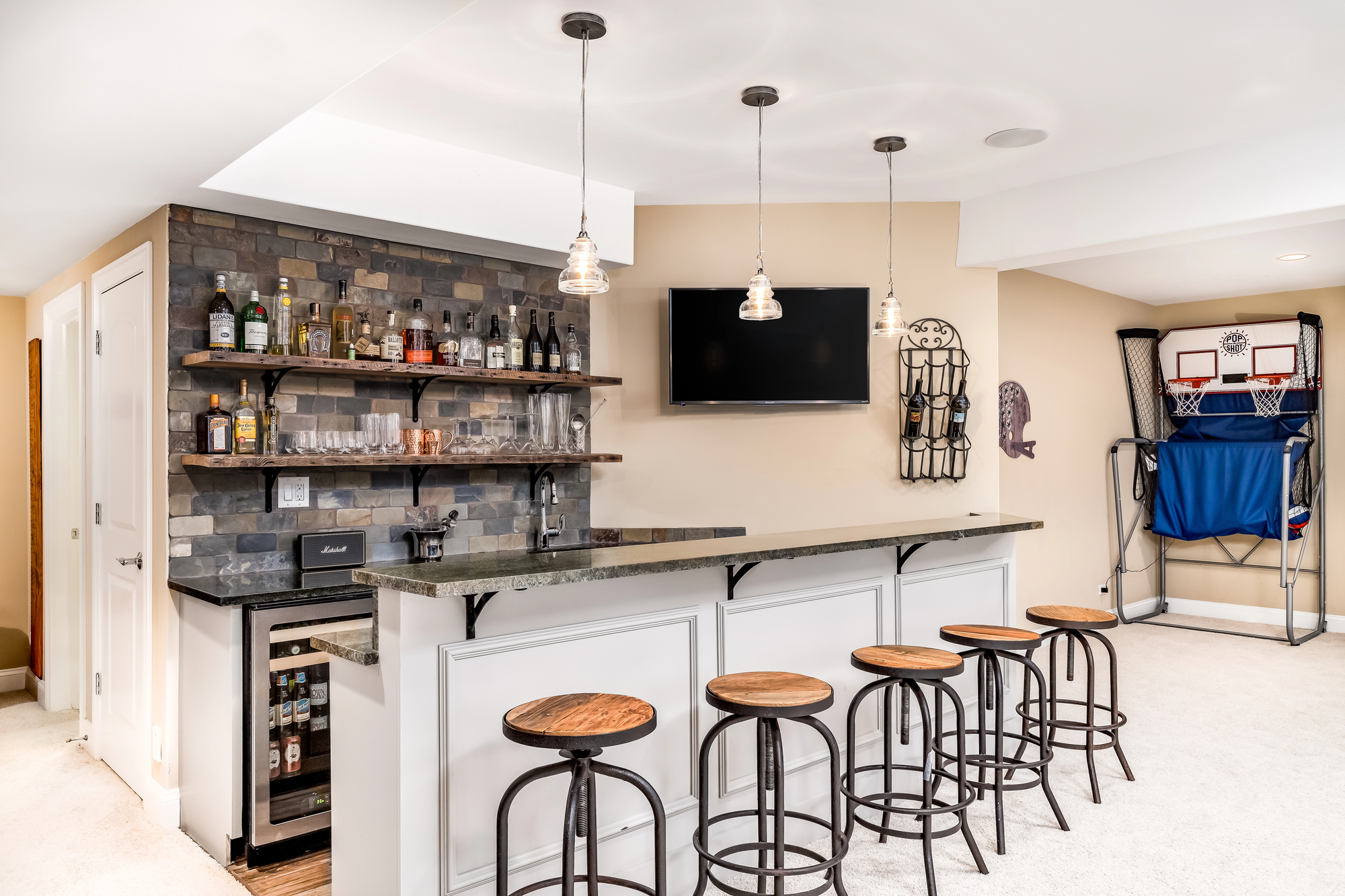 Modern home bar with four wooden stools, a stone backsplash, wall-mounted TV, assorted drinks, and a basketball arcade game in a carpeted room.
