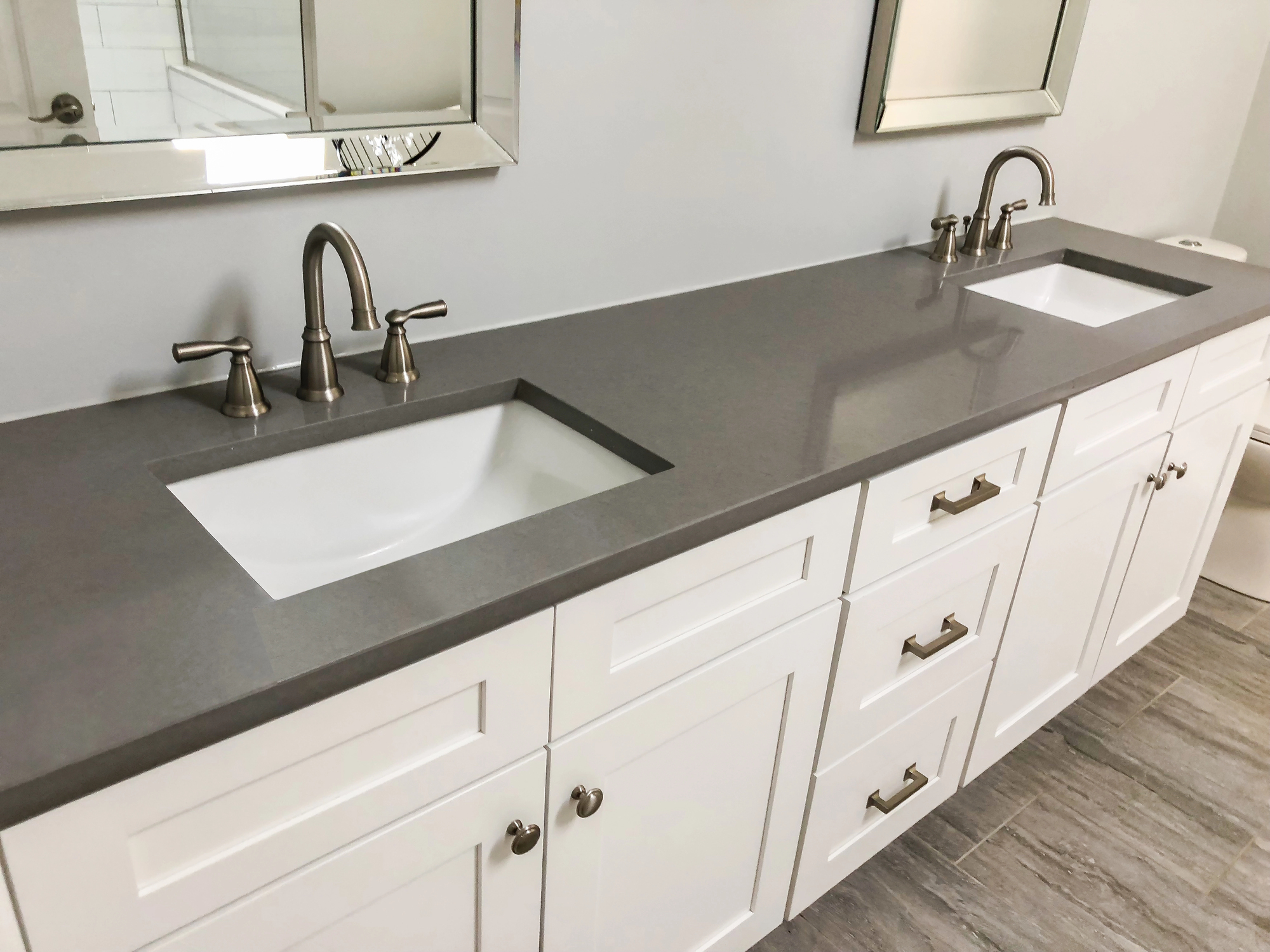 Modern double bathroom vanity with gray countertop, two undermount sinks, chrome faucets, white cabinets, and rectangular mirrors above each sink.