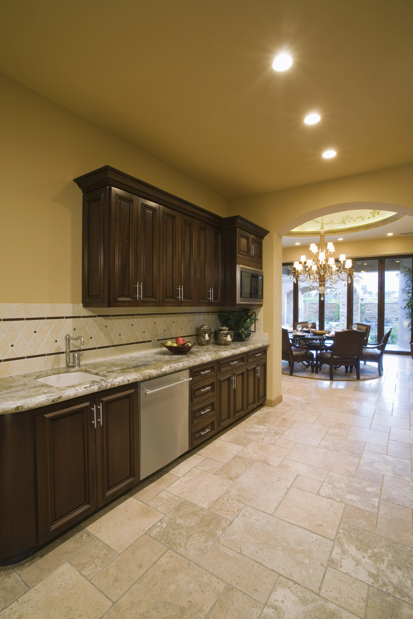 Modern kitchen with dark wood cabinets, granite countertops, beige tile floor, and stainless steel appliances, leading to a dining area with a chandelier and large windows.