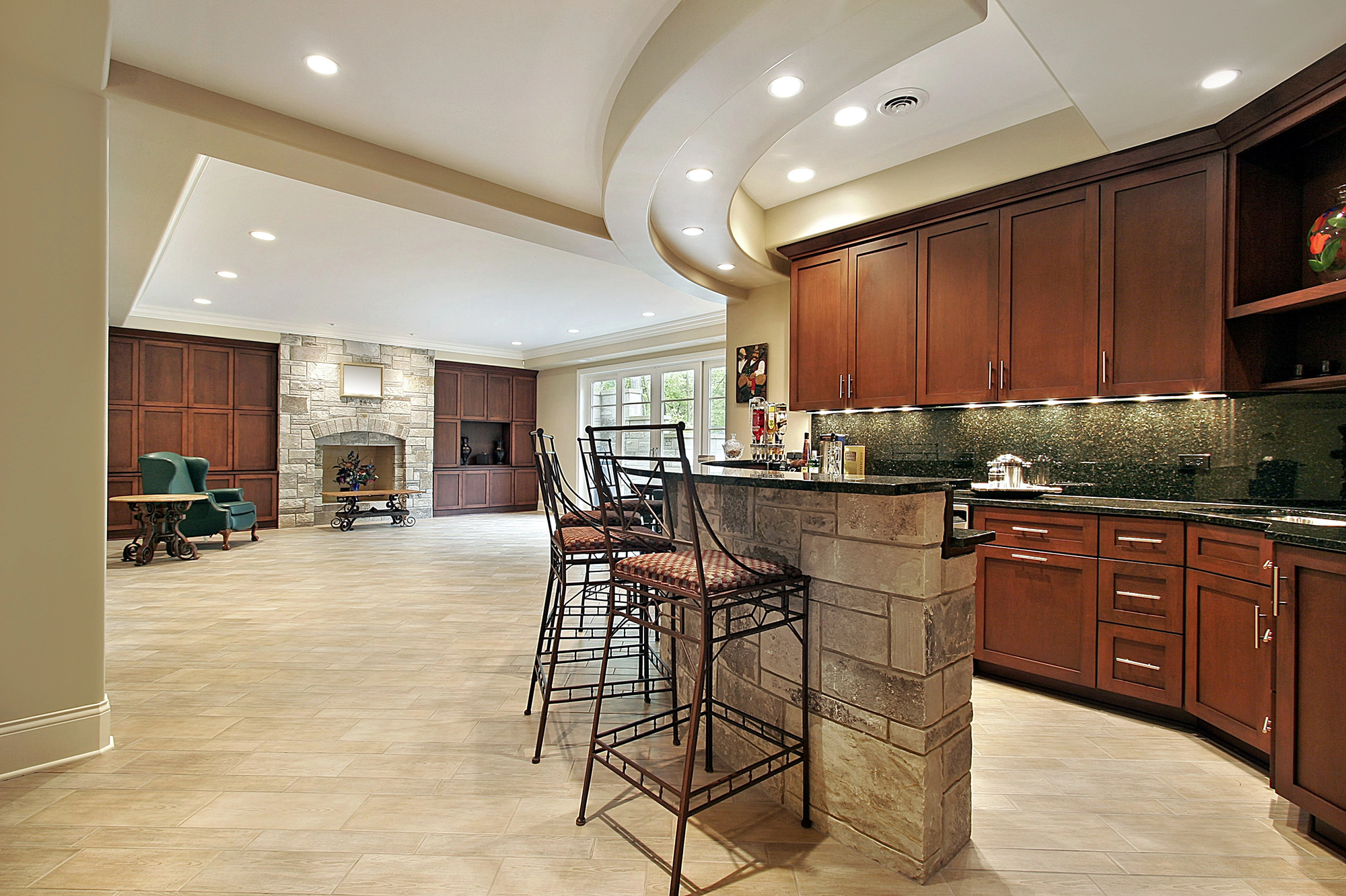 Spacious modern kitchen with dark wood cabinets, a stone breakfast bar with high chairs, and open layout leading to a living area with a stone fireplace and large windows.
