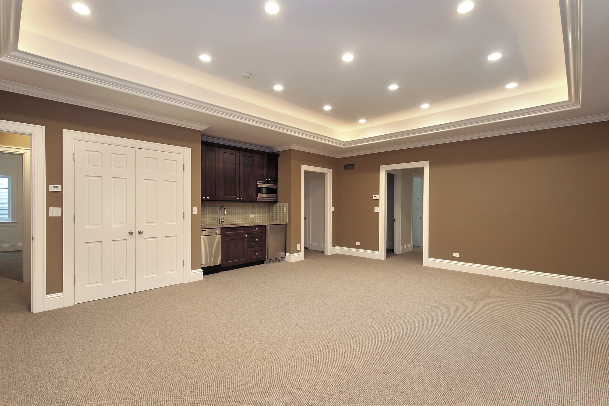 Spacious empty room with beige carpet, brown walls, recessed lighting, a small kitchenette, double doors, and three doorways leading to other rooms.